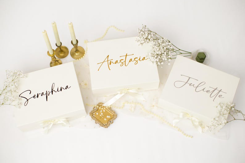 May include: Three square white gift boxes with ribbon closures, each personalised with a name in script lettering: Seraphina, Anastasia, and Juliette. The boxes are arranged with three thin cream candles in gold holders and sprigs of baby's breath.