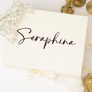 May include: A rectangular white gift box featuring the name Seraphina printed in black script lettering on the lid. The box is adorned with a small white ribbon bow at the bottom edge and is decorated with a sprig of white baby's breath flowers.
