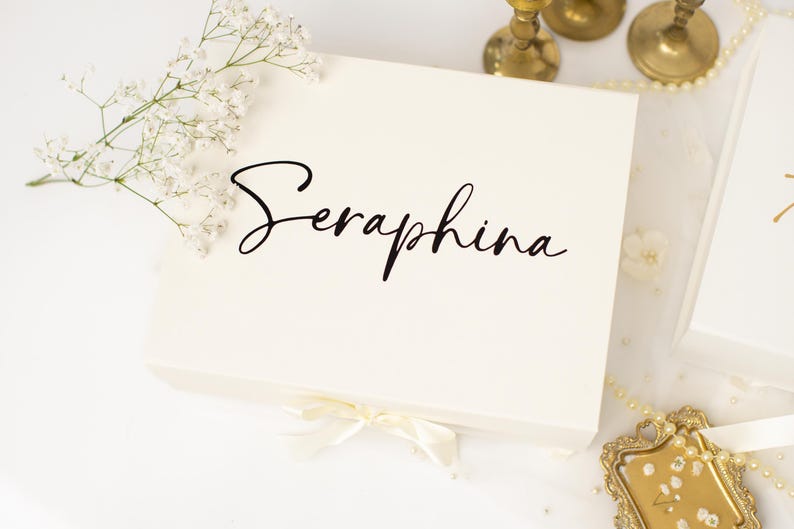 May include: A rectangular white gift box featuring the name Seraphina printed in black script lettering on the lid. The box is adorned with a small white ribbon bow at the bottom edge and is decorated with a sprig of white baby's breath flowers.