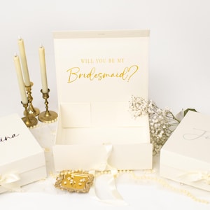 May include: Three cream-coloured rectangular gift boxes with ribbon closures. Two boxes are closed and personalised with the names Seraphina and Julietta in black script. The centre box is open, displaying the gold text Will You Be My Bridesmaid?