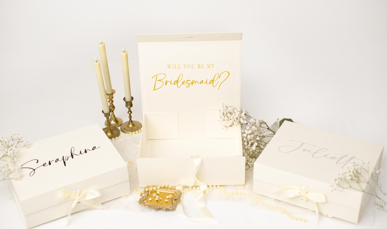 May include: Three cream-coloured rectangular gift boxes with ribbon closures. Two boxes are closed and personalised with the names Seraphina and Julietta in black script. The centre box is open, displaying the gold text Will You Be My Bridesmaid?