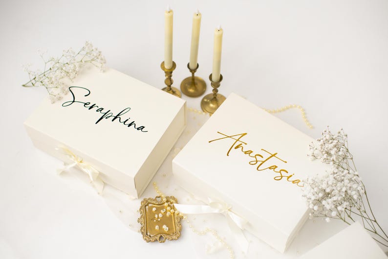 May include: Two ivory gift boxes, one with "Seraphina" in black script and the other with "Anastasia" in gold script. The boxes are tied with ivory ribbons and decorated with white flowers and pearls. Three lit candles in gold holders are in the background.