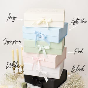 May include: A stack of six rectangular gift boxes with ribbon closures, labelled BOX COLOR OPTIONS. Colours include ivory, light blue, sage green, pink, white, and black. Each box features a matching ribbon tie. Text notes that ribbon colour is fixed.