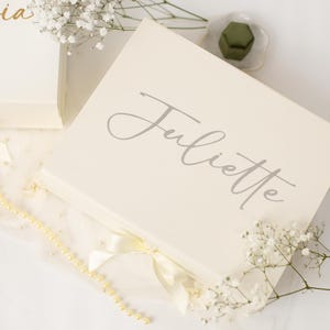 May include: A rectangular cream-coloured gift box featuring the name Juliette in elegant grey script lettering on the lid. The box is accented with a cream satin ribbon bow at the front and is decorated with small white baby's breath flowers.