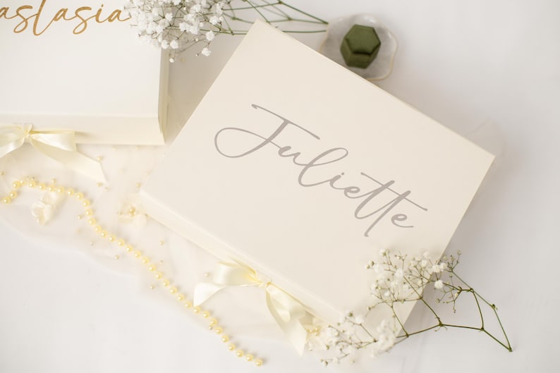 May include: A rectangular cream-coloured gift box featuring the name Juliette in elegant grey script lettering on the lid. The box is accented with a cream satin ribbon bow at the front and is decorated with small white baby's breath flowers.