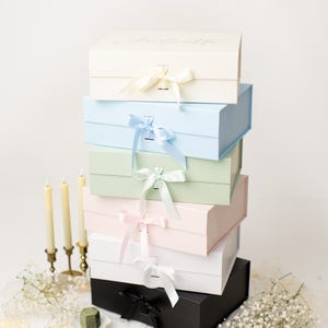 May include: A vertical stack of six rectangular gift boxes in cream, light blue, sage green, pale pink, white, and black. Each box features a matching satin ribbon bow closure at the front. The top cream box has the name Juliette printed in script.