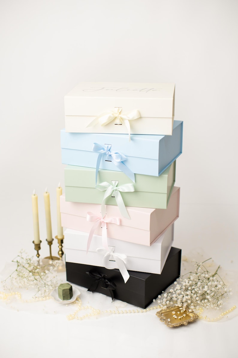 May include: A vertical stack of six rectangular gift boxes in cream, light blue, sage green, pale pink, white, and black. Each box features a matching satin ribbon bow closure at the front. The top cream box has the name Juliette printed in script.