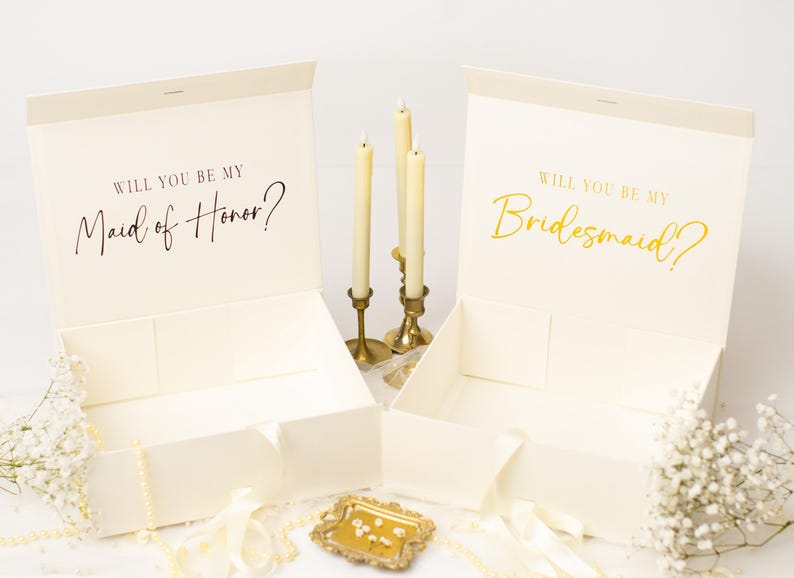 May include: Two open ivory gift boxes with the text "WILL YOU BE MY Maid of Honor?" and "WILL YOU BE MY Bridesmaid?" in elegant script. The boxes are decorated with flowers, candles, and a small decorative tray.