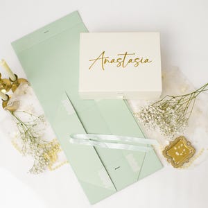May include: A white rectangular gift box with the name Anastasia written in gold script, accompanied by a light green flat-folding box, three thin candles in gold holders, sprigs of baby's breath, and a small gold-framed mirror.