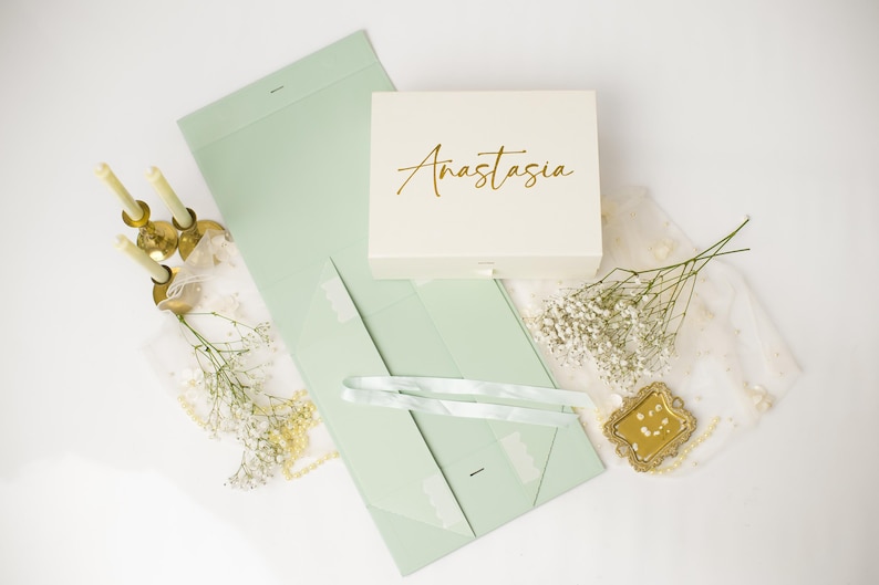 May include: A white rectangular gift box with the name Anastasia written in gold script, accompanied by a light green flat-folding box, three thin candles in gold holders, sprigs of baby's breath, and a small gold-framed mirror.