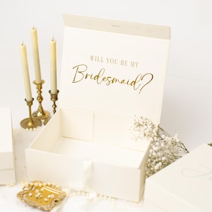 May include: An open cream-coloured rectangular gift box with a magnetic closure. The inside lid features the gold foil text "WILL YOU BE MY Bridesmaid?". The box is empty, with a white ribbon pull tab on the front edge, and has a smooth, matte finish.