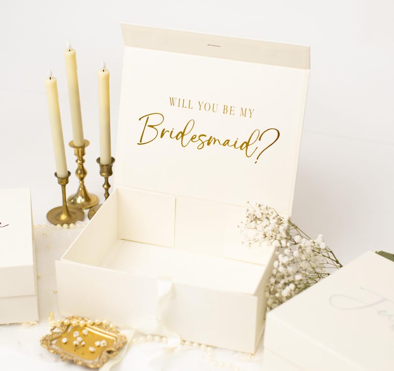 May include: An open cream-coloured rectangular gift box with a magnetic closure. The inside lid features the gold foil text "WILL YOU BE MY Bridesmaid?". The box is empty, with a white ribbon pull tab on the front edge, and has a smooth, matte finish.