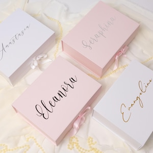 May include: Four rectangular gift boxes with magnetic closures and satin ribbon ties. Two boxes are white and two are light pink. Each box features a custom name in cursive script: Anastasia in black, Seraphina in silver, Eleanora in black, and Evangeline in gold.