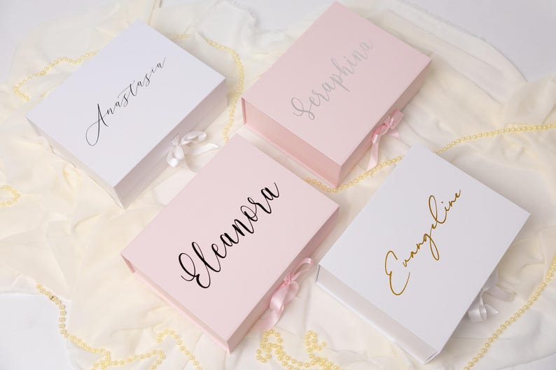 May include: Four rectangular gift boxes with magnetic closures and satin ribbon ties. Two boxes are white and two are light pink. Each box features a custom name in cursive script: Anastasia in black, Seraphina in silver, Eleanora in black, and Evangeline in gold.