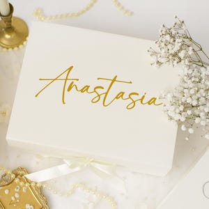 May include: A rectangular white gift box featuring the name Anastasia written in gold script lettering on the lid. The box includes a white ribbon closure at the front and is decorated with a sprig of white baby's breath flowers placed on top.