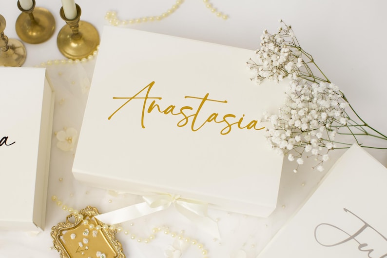 May include: A rectangular white gift box featuring the name Anastasia written in gold script lettering on the lid. The box includes a white ribbon closure at the front and is decorated with a sprig of white baby's breath flowers placed on top.
