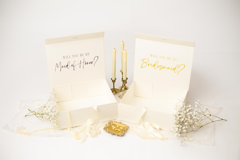 May include: Two open cream-coloured rectangular gift boxes with magnetic lids. The left box features the black text Will You Be My Maid of Honour? and the right box features the gold text Will You Be My Bridesmaid? accompanied by candles and flowers.