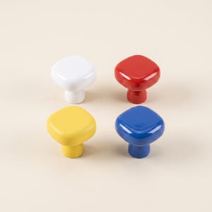 May include: A set of four square-shaped cabinet knobs with rounded corners, featuring a smooth, glossy finish in solid white, red, yellow, and blue colors. Each knob has a central cylindrical base that tapers slightly toward the bottom.