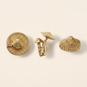 May include: Three gold-toned metal decorative knobs. The first is a circular knob with a concentric spiral pattern and a small rose detail. The second is a dual-mushroom shaped knob. The third is a fan-shaped seashell knob with vertical ridges.