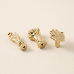 Può includere: Tre pomelli per armadietti in metallo color oro a forma di mani umane. Un pomello è un palmo aperto, uno è un pugno chiuso e uno è un palmo piatto con una superficie testurizzata. Ogni pomello presenta un foro filettato alla base.