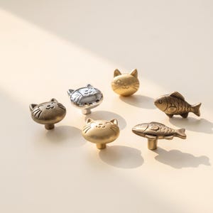 May include: A collection of six cabinet knobs shaped like cats and fish. Three knobs are cat faces in brass and silver tones, while two are fish shapes in a brass finish. Each knob features a central post for mounting to furniture or cabinetry.