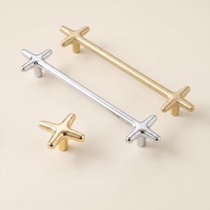 May include: A collection of cabinet hardware featuring two long bar pulls and one single knob. The hardware has a cross-shaped design at each end. One bar pull and the knob have a polished gold finish, while the other bar pull has a polished silver finish.