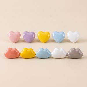 May include: A collection of ten small, smooth, heart-shaped ceramic objects arranged in two rows. The top row features pink, lavender, yellow, light blue, and white hearts. The bottom row includes peach, yellow, light blue, white, and grey hearts.