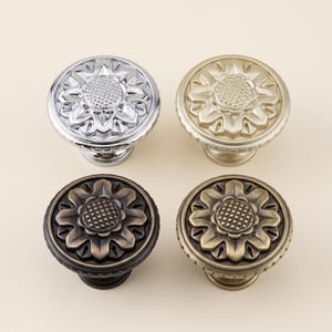 May include: Four round metal cabinet knobs arranged in a square grid. Each knob features a raised floral design with a textured center and petal-like edges. The finishes include polished chrome, brushed gold, dark bronze, and antique brass tones.