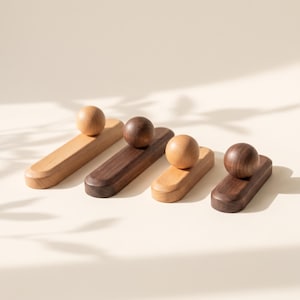 Puede incluir: Cuatro objetos de madera, cada uno compuesto por una base rectangular lisa con extremos redondeados y un pomo esférico fijado en la parte superior. Dos bases y pomos son de madera clara, mientras que los otros dos son de madera oscura.