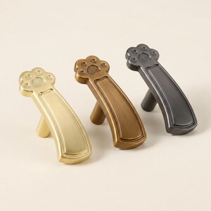 Può includere: Tre pomelli per mobili in metallo a forma di zampa di animale, ciascuno con un manico curvo e un design a cuscinetto testurizzato. Il set include una finitura in oro spazzolato, bronzo antico e nero opaco su un supporto cilindrico.