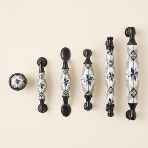 May include: A collection of six cabinet hardware pieces including one round knob and five handles of varying lengths. Each piece features a matte black metal base and a ceramic centre section decorated with a blue, white, and gold star pattern.
