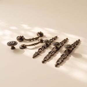 May include: A collection of antique brass-toned metal hardware including one round knob, two curved bail pulls, and three elongated decorative drawer pulls with intricate scrollwork and floral-inspired detailing on the circular centers.