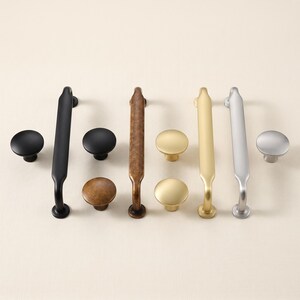 May include: A collection of cabinet hardware including four long, slender drawer pulls and four round knobs. The hardware is finished in matte black, antique bronze, brushed gold, and brushed silver, showcasing a variety of metallic textures.