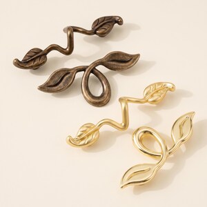 May include: Four metal cabinet pulls shaped like winding vines with leaves. Two pulls have a dark, antique bronze finish, and two pulls have a bright, polished gold finish. Each pull features a curved, organic stem design with detailed leaves.