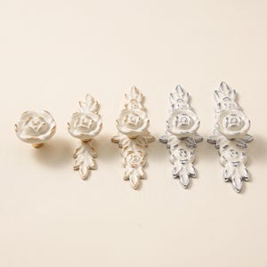 Puede incluir: Un conjunto de cinco pomos y tiradores de armario blancos con un diseño de rosa en relieve y bordes dorados. El set incluye un pomo redondo y cuatro tiradores alargados de distintas longitudes, decorados con hojas esculpidas y motivos florales.