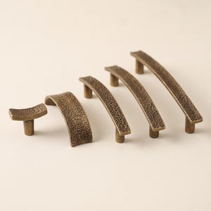 May include: A set of five bronze-toned metal cabinet pulls and a knob, each featuring a distinct hammered texture. The hardware pieces vary in length and curvature, showcasing a dark, aged finish with a consistent metallic sheen throughout.