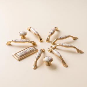 May include: A collection of gold-toned metal cabinet hardware including various drawer pulls and knobs. Each piece features a white ceramic inlay decorated with a delicate floral pattern of small purple and blue flowers with green stems.