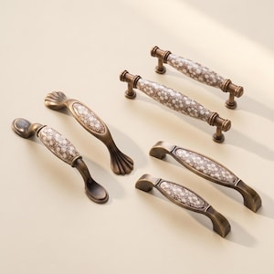 May include: A collection of five cabinet handles featuring a bronze-toned metal finish and central ceramic inserts decorated with a repeating white and brown floral pattern. The handles vary in shape, including curved, arched, and straight designs.