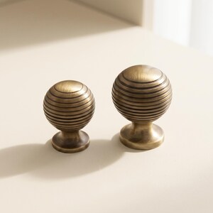 May include: Two spherical cabinet knobs with a brushed antique brass finish. Each knob features a series of horizontal, recessed circular grooves wrapping around the globe-shaped head, supported by a smooth, flared circular base pedestal.