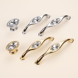 May include: A collection of cabinet hardware including two oval-shaped knobs and four curved handles. The hardware features a polished silver or gold finish with a central, faceted clear crystal inlay in a marquise shape on each piece.