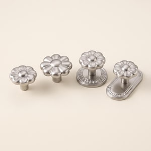 May include: Four silver-toned metal cabinet knobs with a flower-shaped head. Two knobs feature a simple stem base, while the other two are mounted on circular and oval backplates, each decorated with a subtle, repeating radial line pattern.