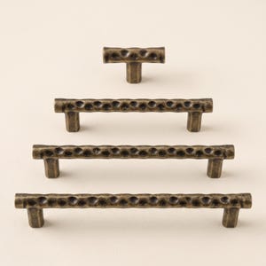 May include: A collection of four bronze-toned metal cabinet pulls and a T-shaped knob arranged in a vertical stack. Each piece features a textured, hammered surface with a series of circular indentations along the horizontal bar.