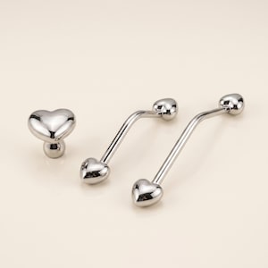 May include: A set of three silver-toned metal body jewelry pieces featuring heart-shaped ends. One piece is a small stud with a heart top, while the other two are curved barbells of different lengths, each finished with a heart at both ends.