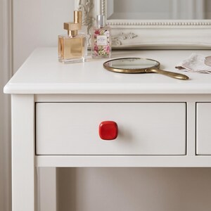 May include: A white wooden vanity table featuring a single drawer with a glossy, square-shaped red knob. The tabletop holds a gold-toned perfume bottle, a small glass perfume bottle with a floral label, and a handheld brass-rimmed mirror.