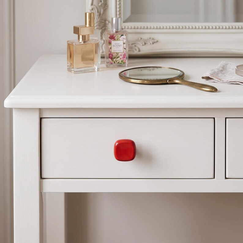 May include: A white wooden vanity table featuring a single drawer with a glossy, square-shaped red knob. The tabletop holds a gold-toned perfume bottle, a small glass perfume bottle with a floral label, and a handheld brass-rimmed mirror.