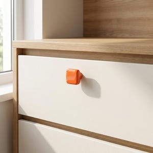 Puede incluir: Un pomo de mueble cuadrado de color naranja brillante con bordes redondeados, montado en un frente de caj&oacute;n de color blanco roto, liso y mate. El pomo presenta un acabado naranja s&oacute;lido y vibrante que contrasta con la superficie neutra.