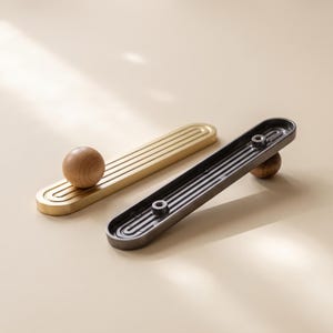 May include: Two elongated cabinet pulls, one in a brushed gold finish and one in a dark charcoal finish, each featuring a central wooden sphere knob and a recessed, ribbed, pill-shaped base with two circular mounting points on the underside.