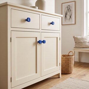 May include: A cream-colored wooden cabinet featuring two top drawers and two lower cabinet doors. The furniture is accented with four square, glossy blue knobs. The cabinet has a clean, flat-panel design with visible hinges on the doors.