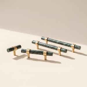 May include: Three cabinet pulls of varying lengths, each featuring a cylindrical dark green marble bar accented with brushed gold metal hardware. The hardware includes circular mounts that secure the stone bars, creating a uniform look.