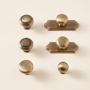 May include: A collection of six cabinet knobs and pulls in a brushed antique brass finish. The set includes two round knobs with concentric ring detailing, two round knobs on rectangular backplates with scalloped edges, and two plain round knobs.